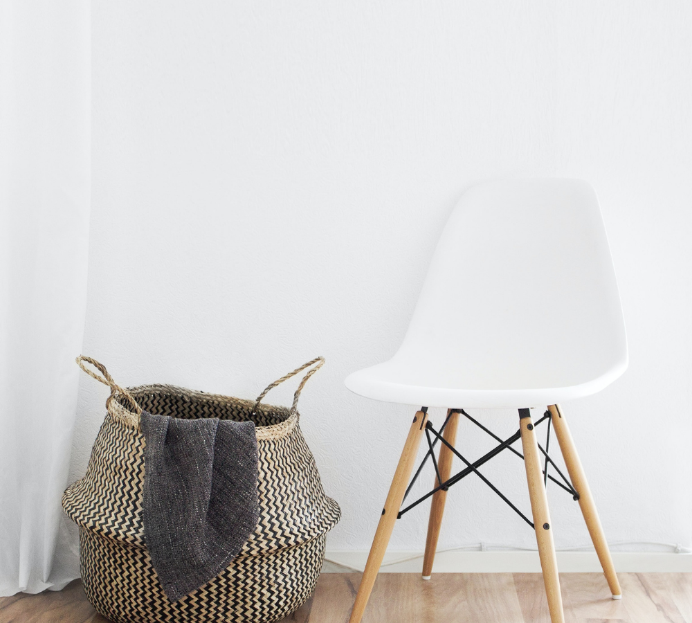 White chair sitting against a white walkk with a brown and tan striped basket and a blanket hanging over the side