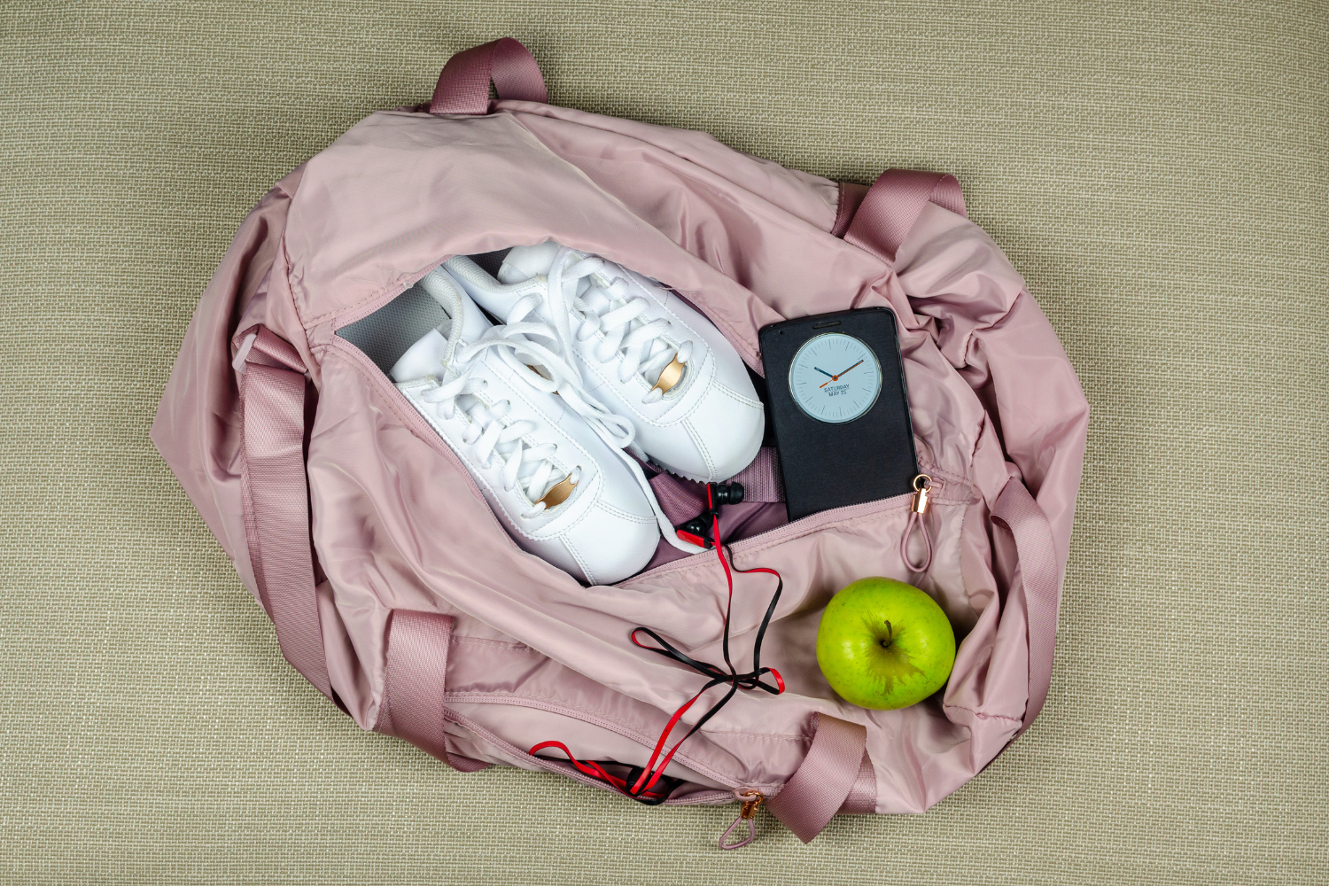 A pink gym bag with a pair of shoes, clock, headphones and a green apple