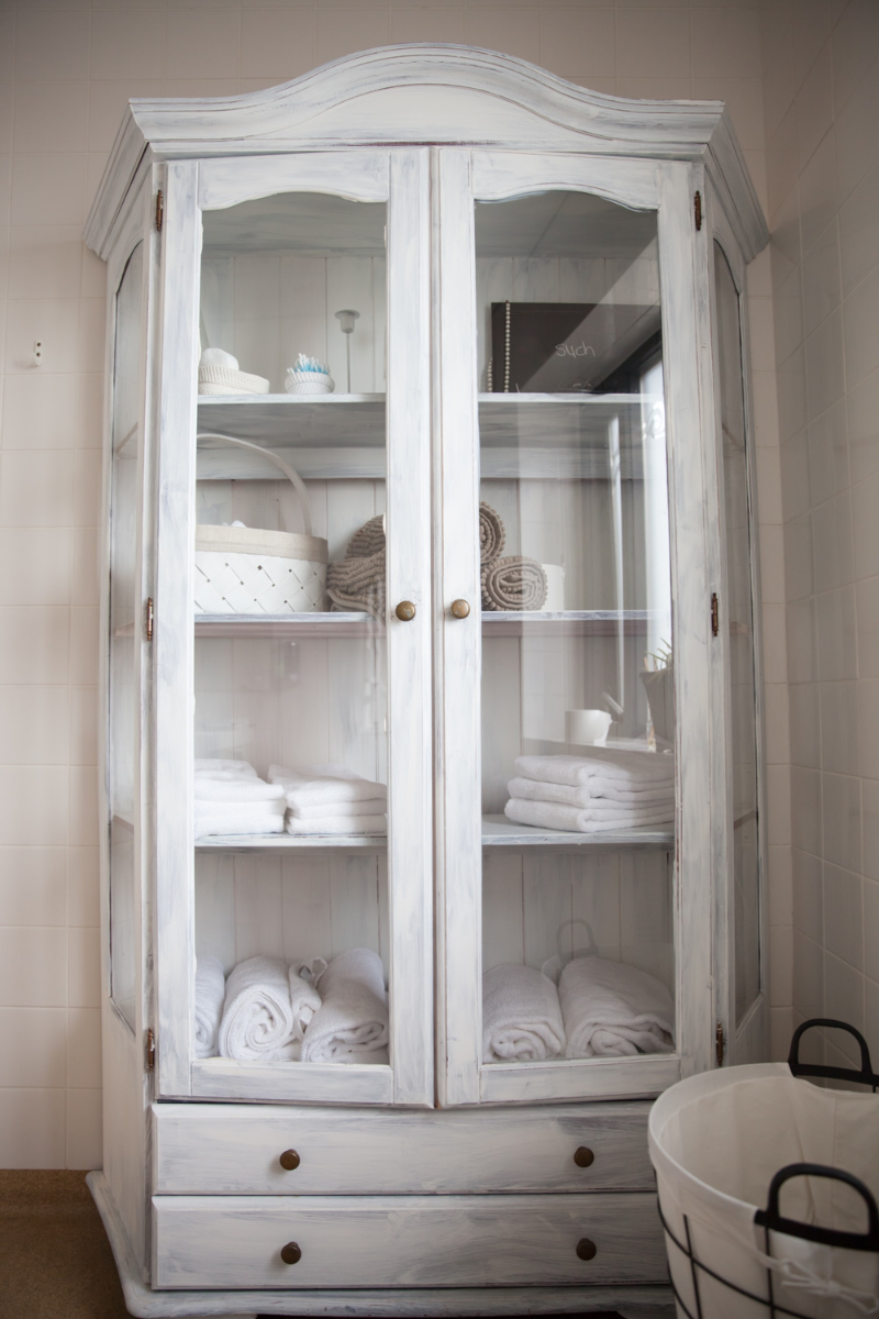 Tall white cabinet with glass windows, full of towels and bathroom decor