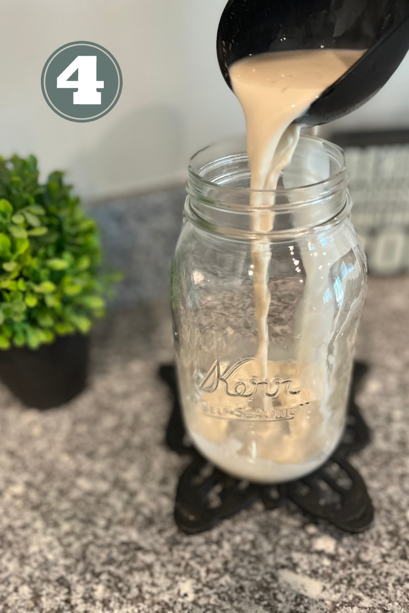 Black spoon pouring creamer into a glass jar on a black trivet. Green plant in the background