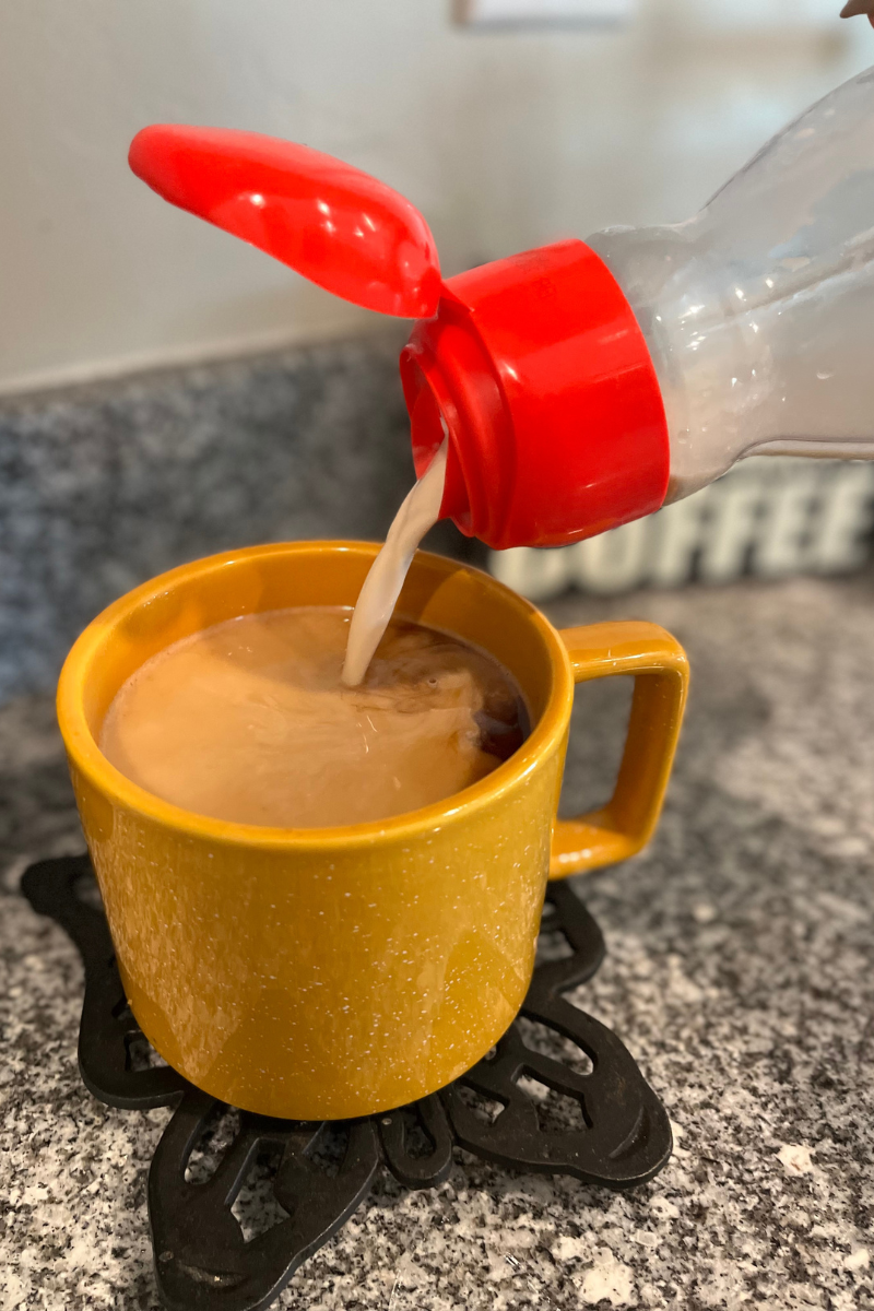 Creamer container with a red top pouring cream into a yellow mug sitting on a black trivet